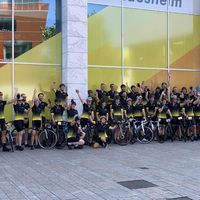 Grande team ciclistico con maglie personalizzate nere e gialle che festeggia con bici davanti a un edificio