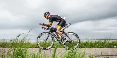 Un triatleta con indosso una tuta da triathlon personalizzata pedala in posizione aerodinamica su una strada costiera durante una prova a cronometro.